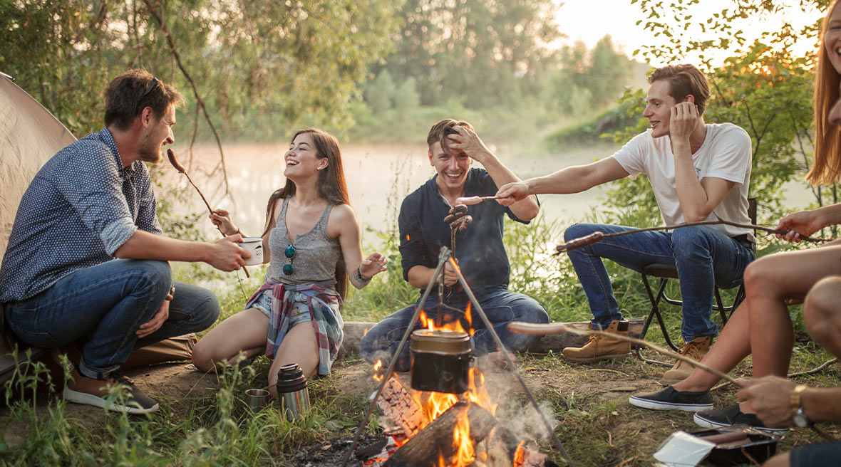 Group of five friends laughing around a campfire in a field surrounded by more trees as the sun sets